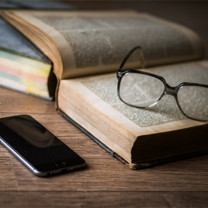 Um óculos sobre um livro e um celular ao lado, ambos em cima de uma mesa de madeira.